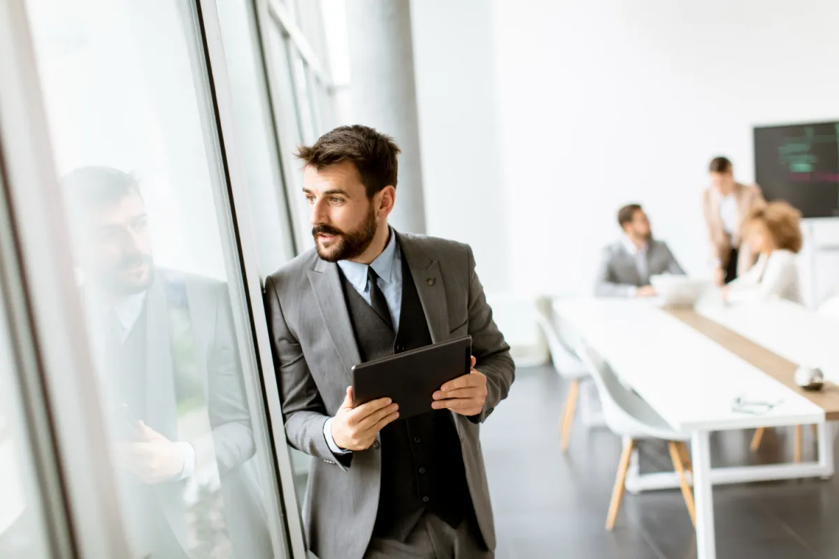 professional man in a suit looking out the window in a bright office ready to hire with his hiring checklist for remote developers on his tablet in his hands