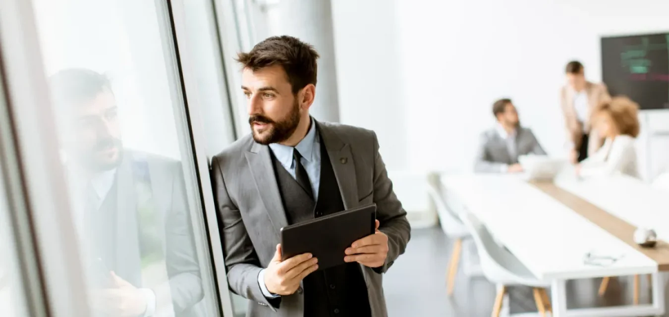 professional man in a suit looking out the window in a bright office ready to hire with his hiring checklist for remote developers on his tablet in his hands